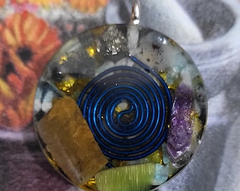 May include: A circular pendant with a silver loop, showcasing a blue spiral design. The pendant is filled with crystals and stones in gold, green, and purple hues. The pendant is set in clear resin.