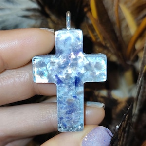 May include: A clear, cross-shaped pendant with blue and white inclusions. The pendant has a silver-colored bail for hanging. The cross is held up against a blurred background of brown and yellow feathers.