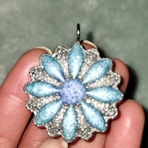 Pode incluir: Pingente prateado em forma de margarida, com pétalas azuis claras e um centro azul mais escuro. O pingente tem uma borda texturizada e ondulada e uma pequena argola para uma corrente.