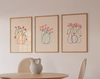 Peut inclure: Trois impressions d'art encadrées présentant des dessins au trait minimalistes de fleurs dans des vases. L'œuvre utilise une palette de couleurs sourdes avec des fleurs orange, roses et violettes. Les cadres sont de couleur marron clair, et le fond est de couleur crème.