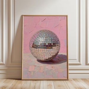 Könnte beinhalten: Eine silberne Discokugel mit Spiegelkacheln liegt auf einer rosa und grauen strukturierten Oberfläche. Die Discokugel ist in einem hellen Holzrahmen gefasst.