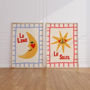 Puede incluir: Dos láminas enmarcadas. Una presenta una luna creciente amarilla con un corazón rojo y las palabras "LA LUNE" en rojo. La otra lámina muestra un sol amarillo con una cara sonriente y las palabras "LE SOLEIL" en rojo.