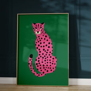 May include: A framed art print featuring a pink cheetah with black spots on a green background. The cheetah is depicted in a seated pose. The frame is gold-colored, and the artwork is displayed against a dark teal wall.