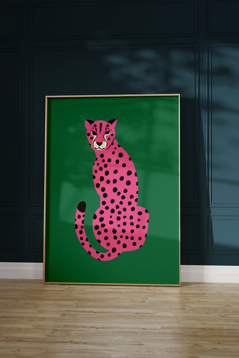 May include: A framed art print featuring a pink cheetah with black spots on a green background. The cheetah is depicted in a seated pose. The frame is gold-colored, and the artwork is displayed against a dark teal wall.