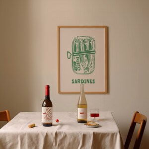 May include: A white tablecloth with a bottle of wine labeled "Le Vin" and a bottle of sparkling wine labeled "BRUT" on a table with a framed print of a sardine can with the text "SARDINES".