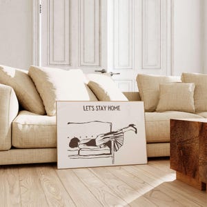 May include: A white framed print with a black and white illustration of a person lying upside down on a couch. The text "Let's Stay Home" is printed at the top of the print.