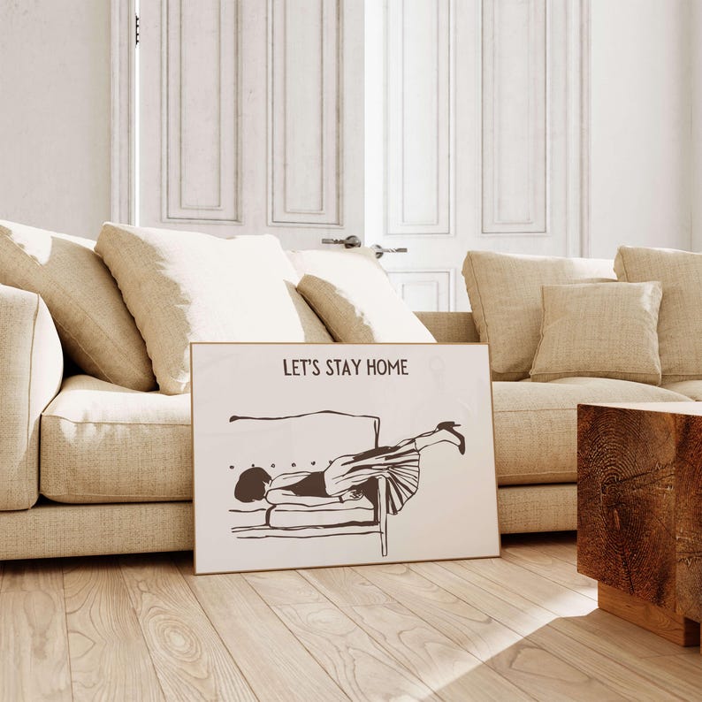 May include: A white framed print with a black and white illustration of a person lying upside down on a couch. The text "Let's Stay Home" is printed at the top of the print.