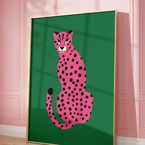 May include: A framed art print featuring a pink cheetah with black spots on a green background. The cheetah is depicted in a seated pose. The frame is gold-colored, and the background is a solid green. The artwork is displayed against a pink wall.
