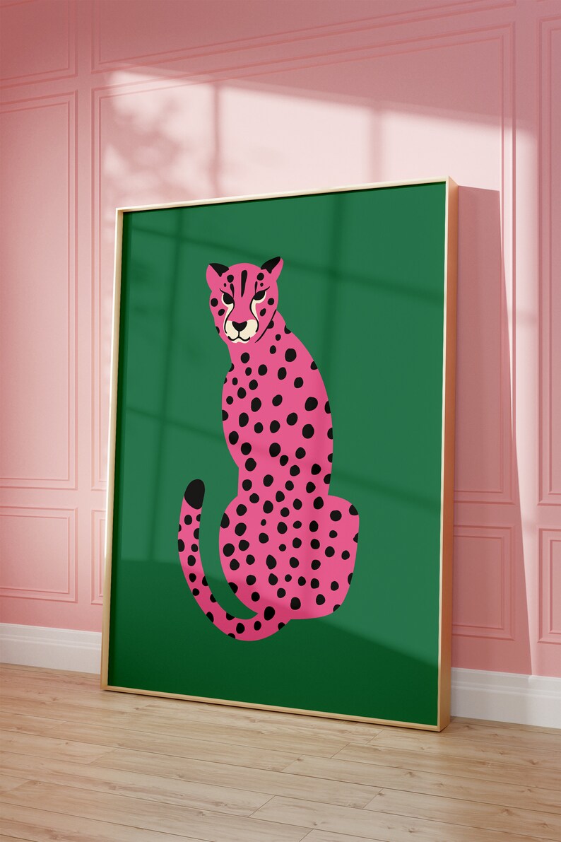 May include: A framed art print featuring a pink cheetah with black spots on a green background. The cheetah is depicted in a seated pose. The frame is gold-colored, and the background is a solid green. The artwork is displayed against a pink wall.