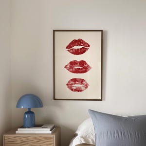 Puede incluir: Una impresi&oacute;n enmarcada con tres marcas de besos de l&aacute;piz labial rojo en una fila vertical sobre un fondo blanco.