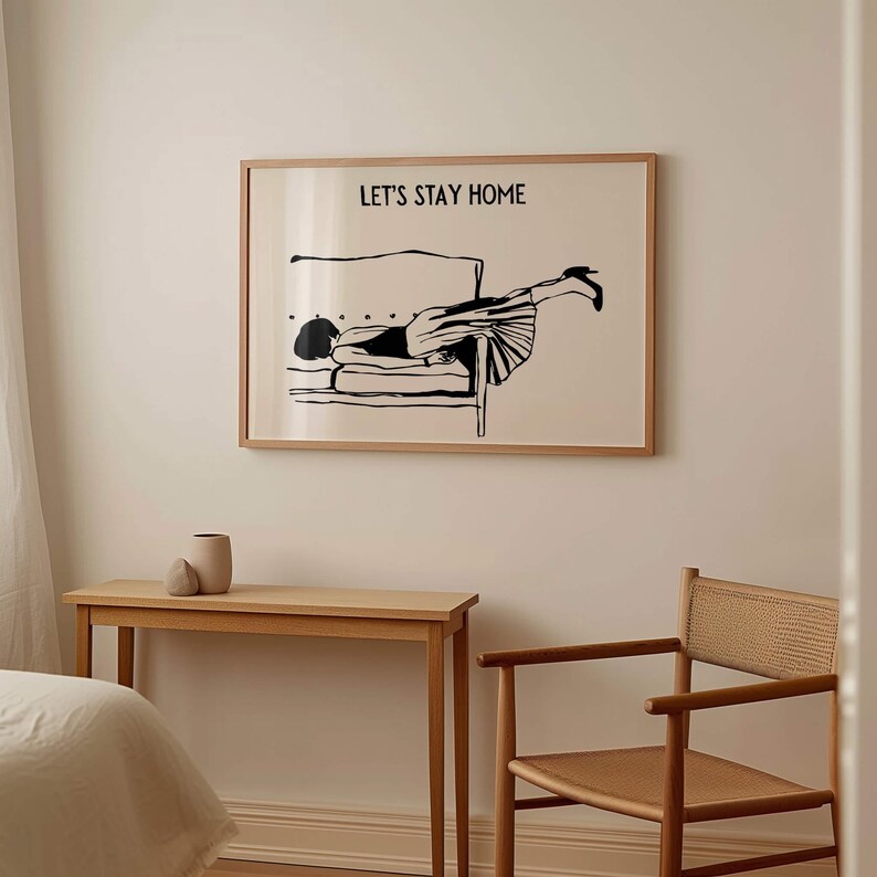 May include: A black and white illustration of a person lying on a couch with the text "Let's Stay Home" above the image. The illustration is framed and hanging on a white wall. A wooden table and chair are in the foreground.