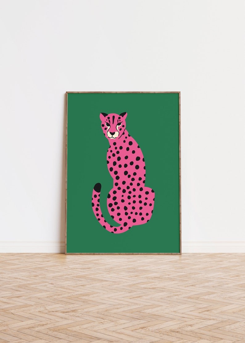 May include: A framed art print featuring a stylized pink cheetah with black spots, set against a vibrant green background. The cheetah is depicted in a seated pose, with a focused expression. The artwork is displayed in a wooden frame.