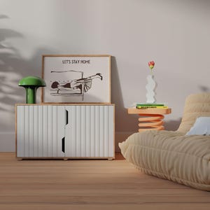 May include: A white cabinet with two doors and a green mushroom lamp on top. A framed print with the text "Let's Stay Home" and a black and white illustration of a woman lying on a couch is above the cabinet. A beige couch with a white pillow is in the background.