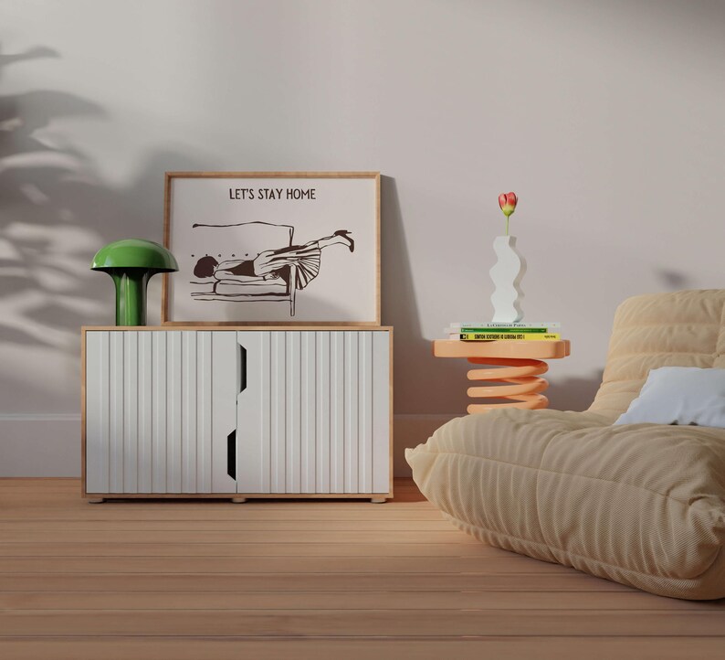 May include: A white cabinet with two doors and a green mushroom lamp on top. A framed print with the text "Let's Stay Home" and a black and white illustration of a woman lying on a couch is above the cabinet. A beige couch with a white pillow is in the background.