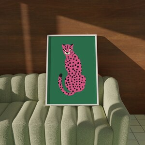 May include: A framed art print featuring a pink cheetah with black spots on a green background. The cheetah is depicted in a seated pose. The artwork is displayed on a light green sofa with a wooden wall in the background.