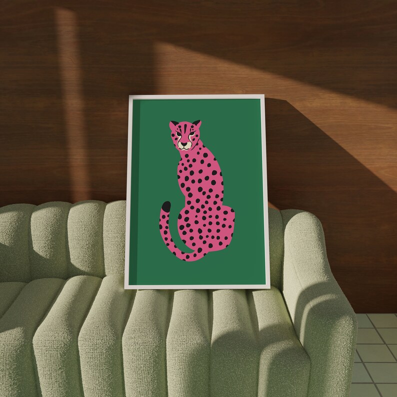May include: A framed art print featuring a pink cheetah with black spots on a green background. The cheetah is depicted in a seated pose. The artwork is displayed on a light green sofa with a wooden wall in the background.