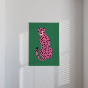 May include: A framed art print featuring a pink cheetah with black spots, set against a vibrant green background. The cheetah is depicted in a seated pose, with its tail curled. The artwork is displayed on a white wall, adding a pop of color to the space.