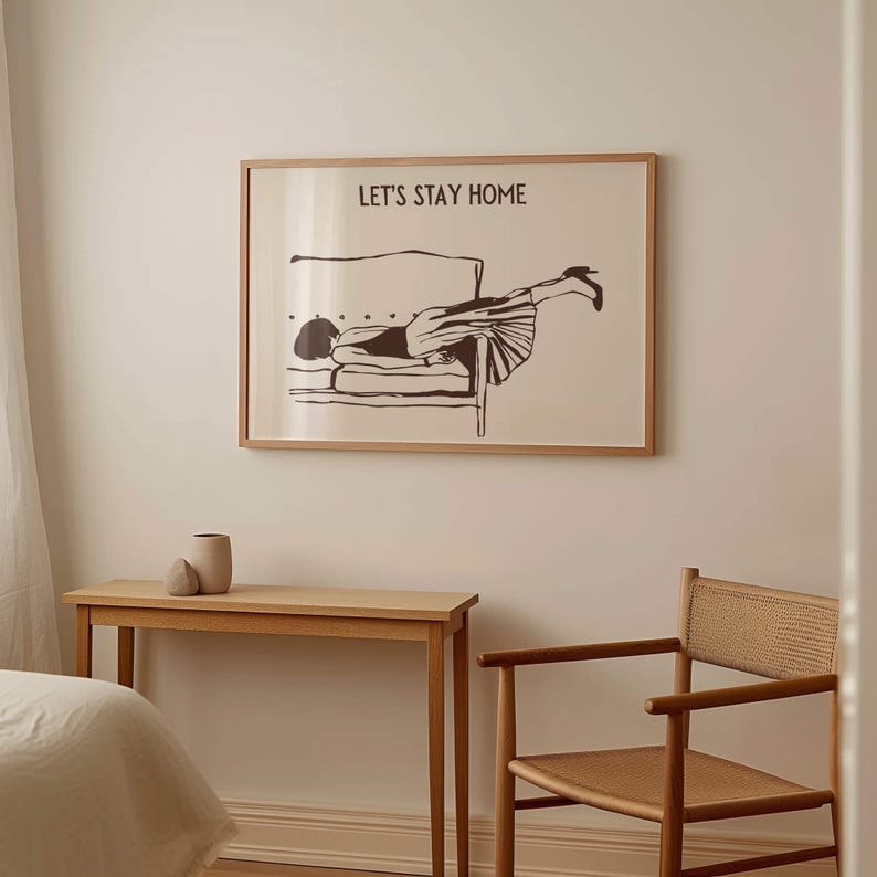 May include: A framed print with a black and white illustration of a woman lying on a couch. The text "LET'S STAY HOME" is printed above the illustration.