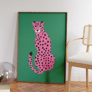 May include: A framed art print featuring a pink cheetah with black spots on a green background. The cheetah is depicted in a seated pose. The artwork is in a wooden frame, suitable for wall decor.