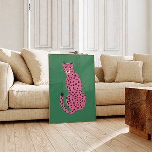 May include: A framed art print featuring a pink cheetah with black spots on a green background. The cheetah is depicted in a seated position. The artwork has a gold frame and is displayed in a living room setting.