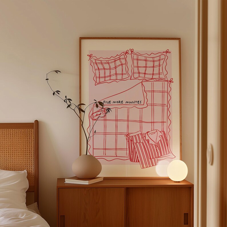 Puede incluir: Una ilustraci&oacute;n enmarcada de una cama a cuadros rosa y blanca con el texto "Five More Minutes" en la manta. La ilustraci&oacute;n est&aacute; en una pared blanca sobre una c&oacute;moda de madera con un jarr&oacute;n y una l&aacute;mpara de globo blanca.