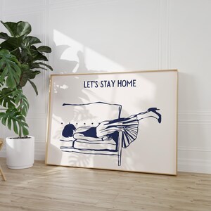 May include: A white framed print with a blue illustration of a person lying on a couch. The text "LET'S STAY HOME" is printed in blue at the top of the print.
