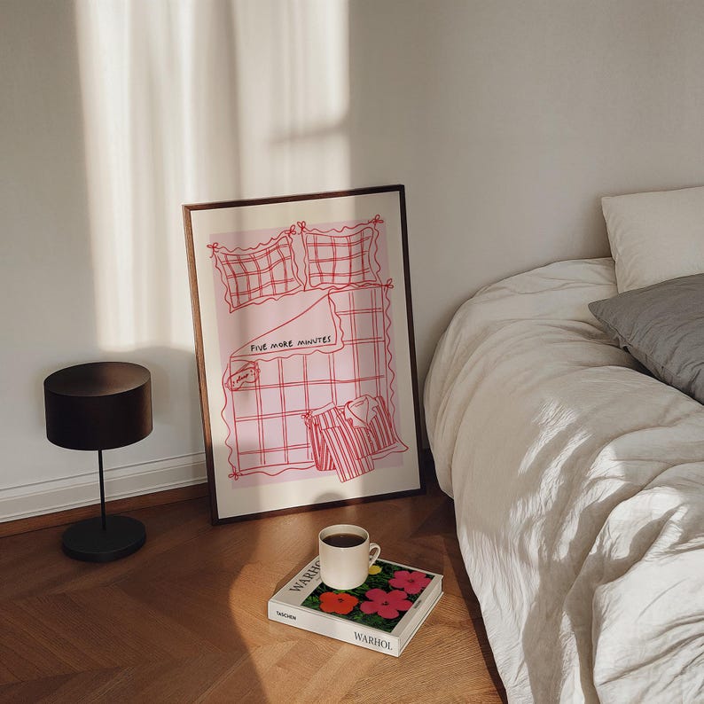 Puede incluir: Una ilustraci&oacute;n enmarcada de una cama con un patr&oacute;n de cuadros rosa y blanco. El texto "Five more minutes" est&aacute; escrito en la cama. Una taza de caf&eacute; y un libro con un dise&ntilde;o de flores est&aacute;n sobre una mesa frente a la ilustraci&oacute;n enmarcada.