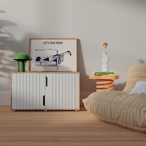May include: A white cabinet with vertical lines and two doors. A framed print with the text "Let's Stay Home" and a black and white illustration of a person lying on a couch. A green lamp with a mushroom-shaped shade is on top of the cabinet. A small orange table with a spring base holds a white vase with a red flower and a stack of books.