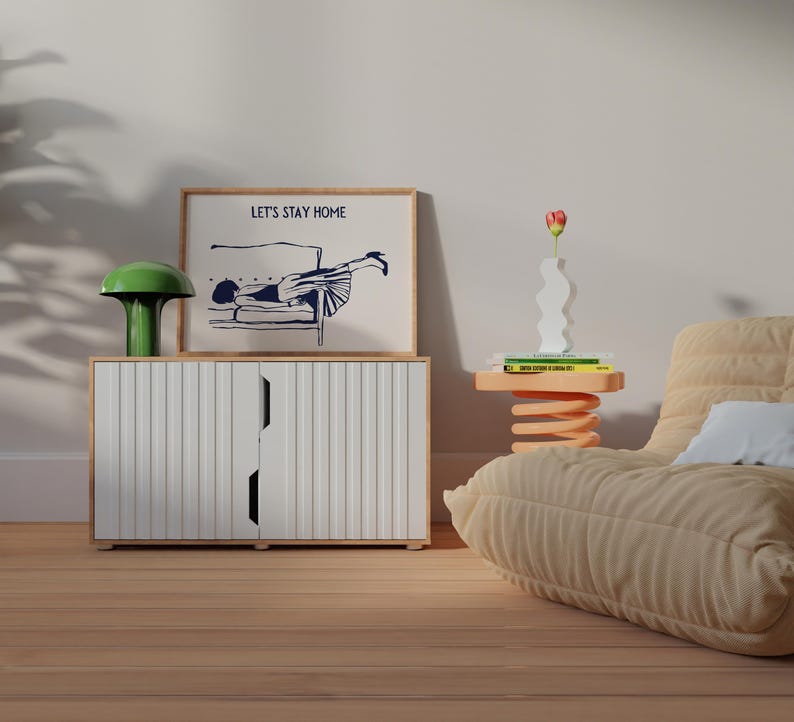 May include: A white cabinet with vertical lines and two doors. A framed print with the text "Let's Stay Home" and a black and white illustration of a person lying on a couch. A green lamp with a mushroom-shaped shade is on top of the cabinet. A small orange table with a spring base holds a white vase with a red flower and a stack of books.