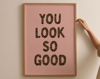 May include: A framed print with the words "YOU LOOK SO GOOD" in a leopard print font on a pink background. The frame is a light brown color. The print is displayed on a white wall.