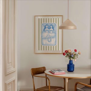 Pu&ograve; includere: Una stampa artistica incorniciata con un'illustrazione blu e bianca di due figure a letto, su uno sfondo a righe verdi e bianche. L'opera &egrave; appesa sopra un tavolo rotondo con un vaso di fiori e una lampada a sospensione.