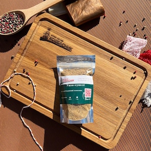 Puede incluir: Una tabla de cortar rectangular de madera con un logotipo grabado, que presenta una bolsa de plástico transparente con especias molidas de color marrón claro etiquetada "Уцхо-сунелі" con un código QR. Una cuchara de madera con granos de pimienta.