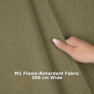 May include: Olive green M1 flame-retardant fabric, 300 cm wide. The material has a textured appearance. A hand is touching the fabric. The fabric is a shade of green.