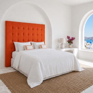 Peut inclure: Une grande tête de lit rectangulaire orange capitonnée avec des boutons, placée derrière un lit recouvert de linge blanc. Deux oreillers blancs présentent de petites bordures orange brodées, et le lit repose sur un tapis marron texturé.