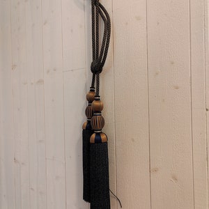 May include: Decorative black and gold curtain tieback with a knotted rope design. The tieback features two decorative tassels with black fringe and gold and navy blue accents. The tassels are attached to a gold and navy blue beaded section.