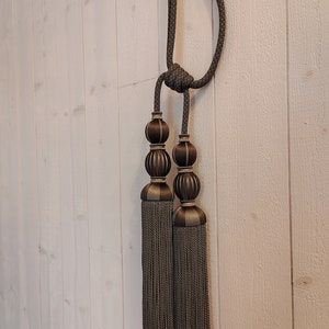 May include: Decorative curtain tiebacks with a dark gray rope and tassels. The tiebacks feature two decorative, ridged spheres in a bronze color. The tassels are a dark gray color, with a matching rope.