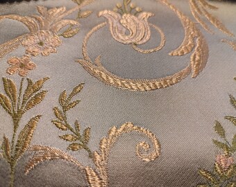 Può includere: Primo piano di un tessuto azzurro con un motivo floreale ornato. Il design presenta fiori, foglie e viticci ricamati in oro e rosa chiaro. Il tessuto ha una leggera lucentezza, suggerendo un possibile utilizzo per tappezzeria o scopi decorativi.
