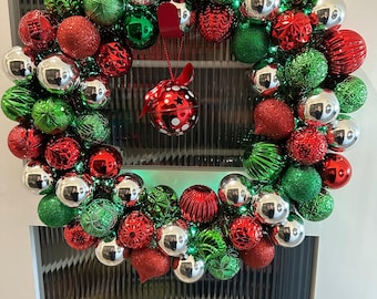 Puede incluir: Una corona navideña festiva compuesta por adornos rojos, verdes y plateados. La corona presenta una variedad de texturas, incluyendo esferas lisas, con purpurina y estampadas. Un pequeño adorno colgante con lunares blancos y un lazo rojo añade un toque decorativo.