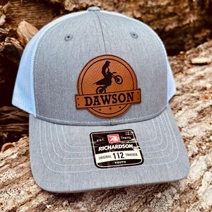 May include: Gray and white youth trucker hat with a brown leather patch featuring a silhouette of a person riding a dirt bike. The patch says "Dawson" in a white banner. The hat is made by Richardson.