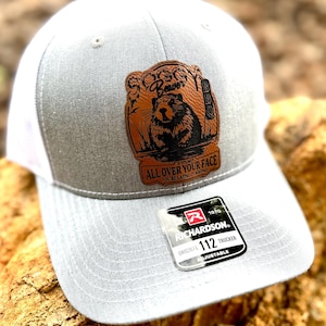 May include: Gray and white trucker hat with a brown leather patch featuring a beaver with the text "Soggy Beaver BBQ" and "It's not all over your face, you're eating it wrong". The hat has a Richardson 112 Trucker label.