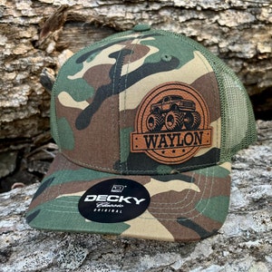 May include: A camouflage trucker hat with a brown leather patch featuring a monster truck and the name "WAYLON" in black lettering. The hat is made by "DECKY Classic Original".