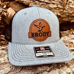 May include: Gray and black youth trucker hat with a brown leather patch featuring a flying duck and the name "Brody". The hat also has a red and white label with the text "Richardson 112 Original Trucker Youth".
