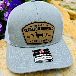 May include: A gray trucker hat with a white mesh back and a curved brim. The hat features a brown leather patch with the text "CLARKSON KENNELS COON HOUNDS" and a dog silhouette. The hat is adjustable and has a Richardson tag.