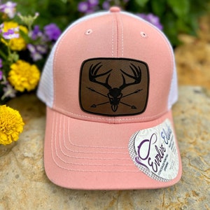 May include: A pink and white trucker hat with a brown leather patch featuring a skull with antlers and crossed arrows. The hat has a white mesh back and a pink front.