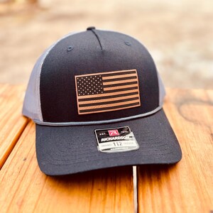 May include: A black and gray trucker hat with a brown leather American flag patch. The hat has an adjustable strap with a black label that reads "Richardson 112 Adjustable".
