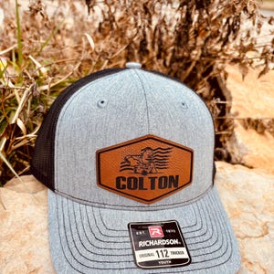 May include: A gray and black trucker hat with a brown leather patch that says "COLTON" and a cartoon image of a person. The hat has a black mesh back and a Richardson 112 Youth label.