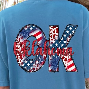 May include: A light blue t-shirt with a red, white, and blue "OK" graphic. The graphic is made up of stars and stripes, and the letters are outlined in black. The word "Oklahoma" is written in red cursive below the graphic.