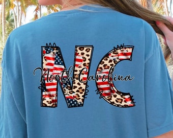 May include: A light blue t-shirt featuring a graphic of the letters "NC" in a patchwork design of an American flag and leopard print. The text "North Carolina" is written below the letters.