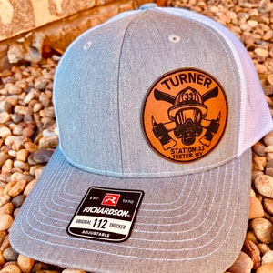 May include: Gray and white trucker hat with a brown leather patch featuring a firefighter helmet, crossed axes, and the text "TURNER 33 STATION 33 TEETER, WY". The hat is adjustable and has a Richardson 112 label.