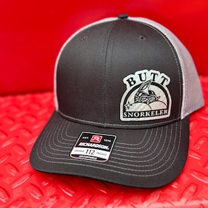 May include: Black and grey trucker hat with a white patch that says "Butt Snorkeler" and a black and white illustration of a person snorkeling. The hat is made by Richardson 112.